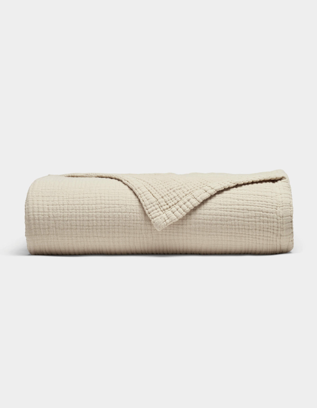 The Cozy Earth Cotton Gauze Blanket, in beige with a soft grid texture, is neatly rolled with a corner folded back to reveal its fabric, shown against a plain white background.