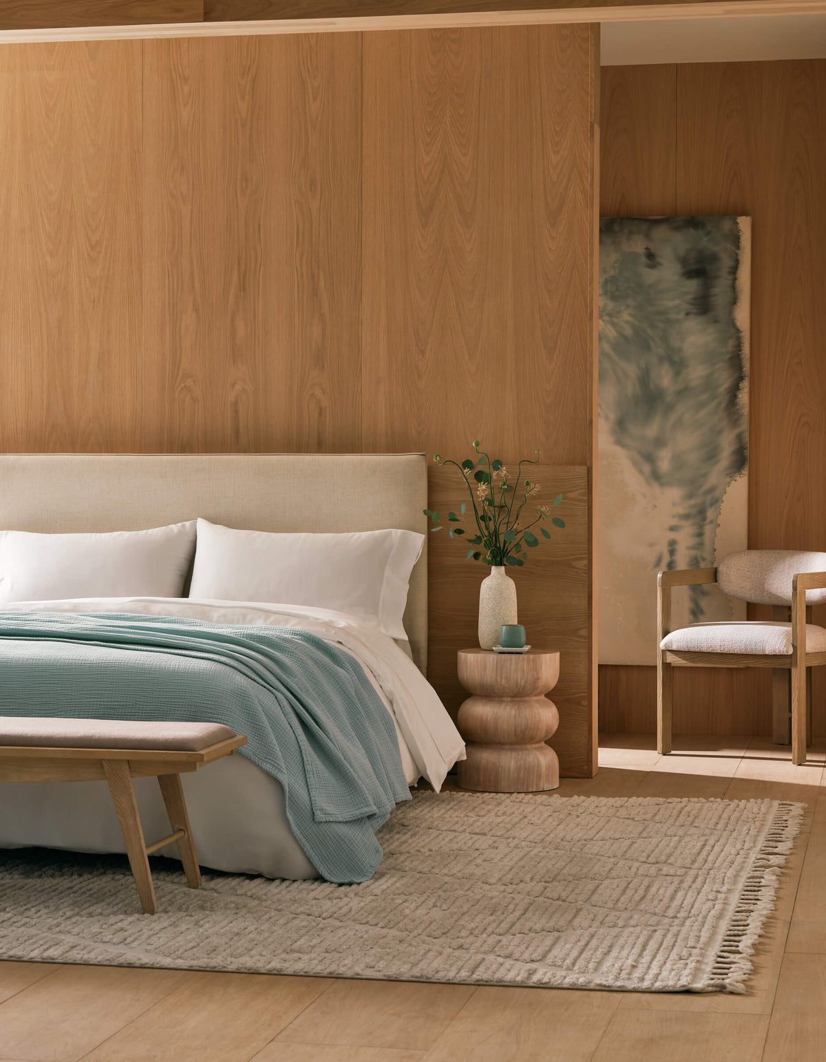 A modern bedroom with light wood-paneled walls, a cream bed topped with Cozy Earth's Cotton Gauze Blanket, white and light blue bedding, a layered side table with greenery, a beige bench, and a soft woven rug.