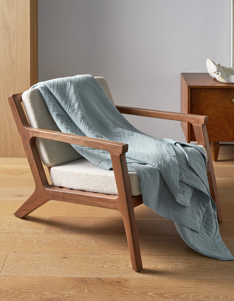 A mid-century modern wooden armchair with cream upholstery sits on a light wood floor, draped with the Cozy Earth Waterfront Grid Matelasse Blanket in light blue. In the background, a wooden nightstand and decorative object are visible.