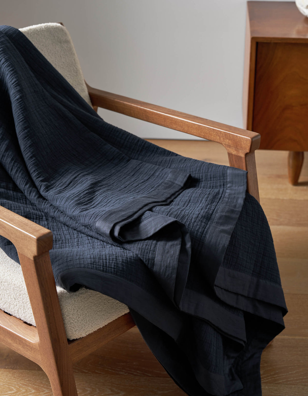 The Cozy Earth Waterfront Grid Matelasse Blanket in dark navy blue is draped over a light upholstered wooden armchair beside a wooden side table on a light wood floor.