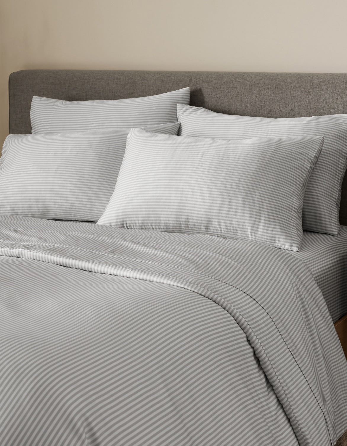 A neatly made bed with a gray upholstered headboard is dressed in light gray and white pinstriped bedding and comforter, with multiple pillows—including Bamboo Pillowcases by Cozy Earth—arranged against a beige wall.|Color:Classic Stripe in Dew