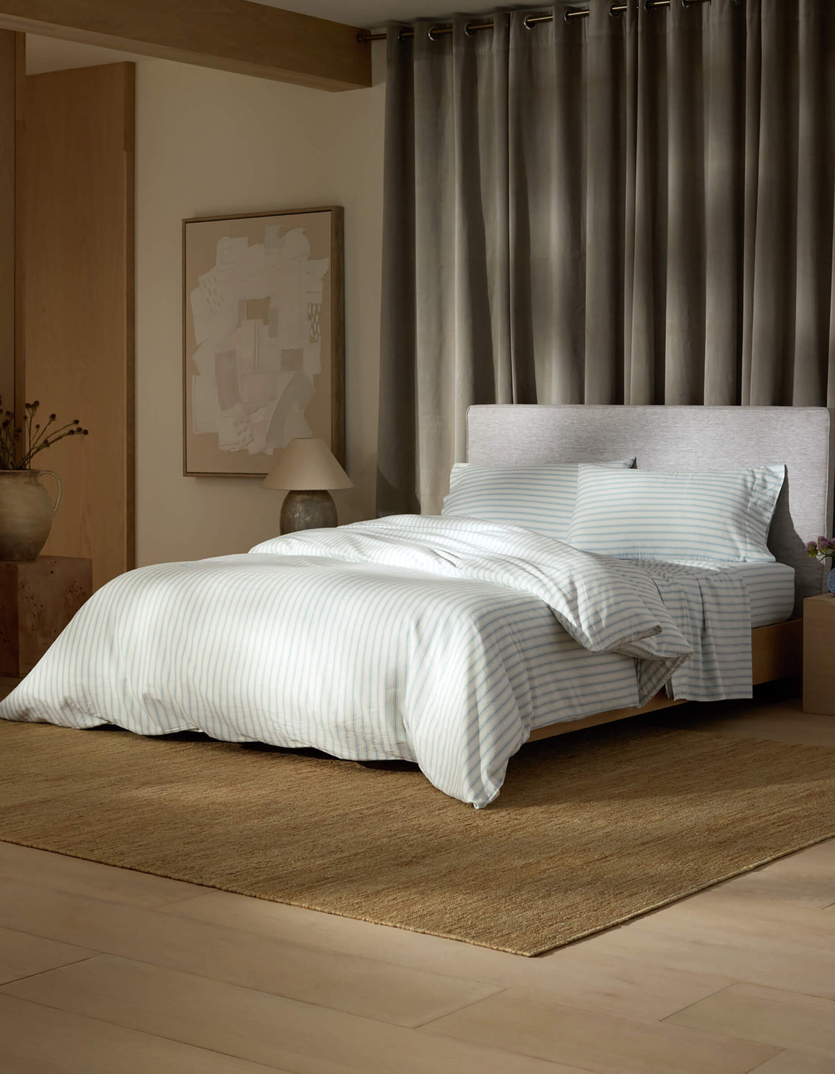 A modern bedroom features the Soft-Wash Cotton Bedding Deluxe Bundle by Cozy Earth in light blue and white stripes, on a brown rug with grey curtains, an abstract art piece, a lamp, and a decorative vase on the side table.