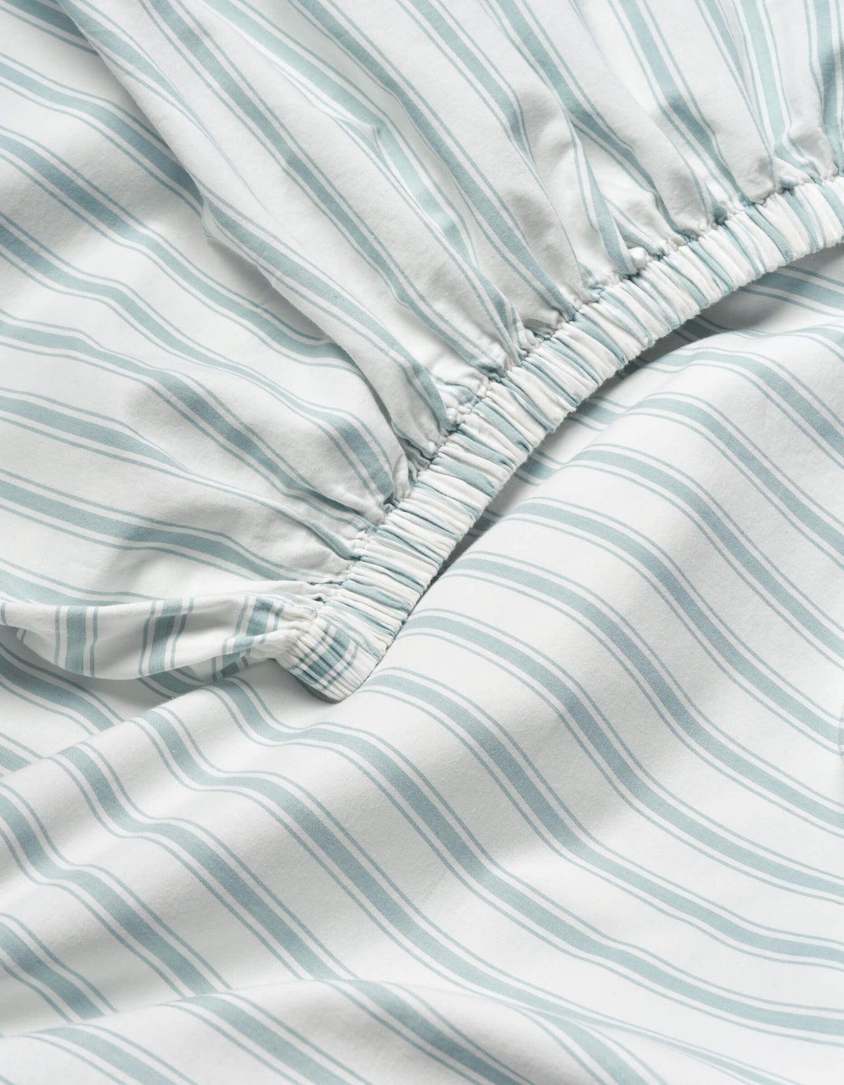 Close-up of the Soft-Wash Cotton Fitted Sheet by Cozy Earth, showing light blue and white vertical stripes and an elastic gathered edge for a snug fit.
