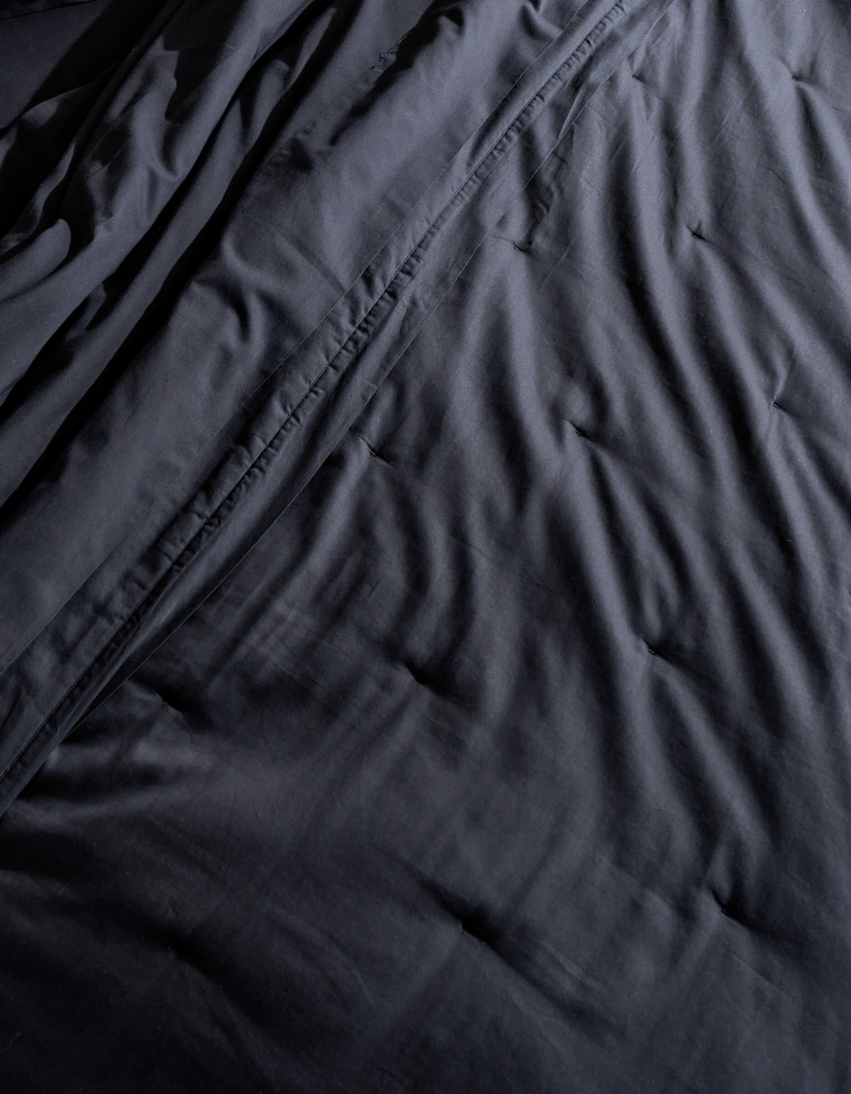 A close-up of Cozy Earth's Soft-Wash Cotton Quilt shows its dark, crumpled fabric with visible wrinkles and folds. The soft lighting highlights the quilt's subtle sheen and textured appearance.
