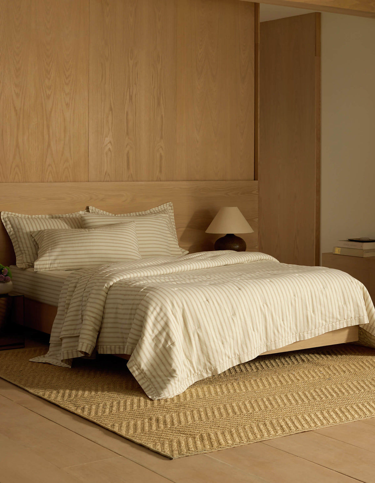 A cozy bedroom featuring Cozy Earth's Soft-Wash Cotton Quilt, cream and beige striped bedding, two pillows, a wooden headboard, a small bedside lamp, a woven rug, and minimalistic wooden furniture in neutral tones.