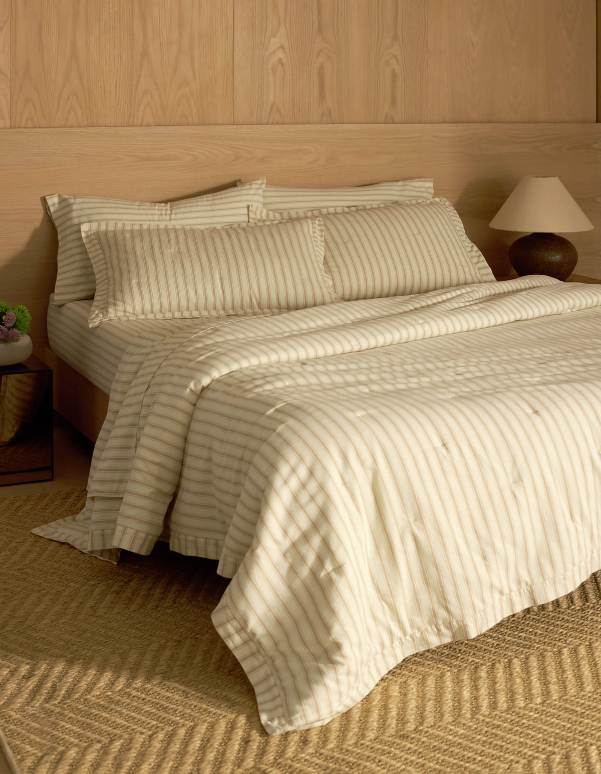 A bed featuring Cozy Earth's Soft-Wash Cotton Quilted Shams sits against a wooden paneled wall, complemented by matching pillows and a comforter. A bedside table with a lamp and vase, plus a woven rug on the floor, complete the cozy scene.