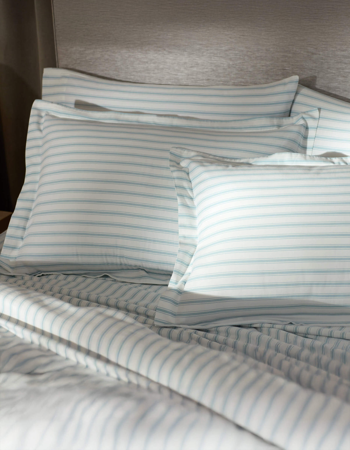 A bed featuring Cozy Earth Soft-Wash Cotton Shams in white and light blue stripes is set against a gray upholstered headboard, softly illuminated by sunlight.