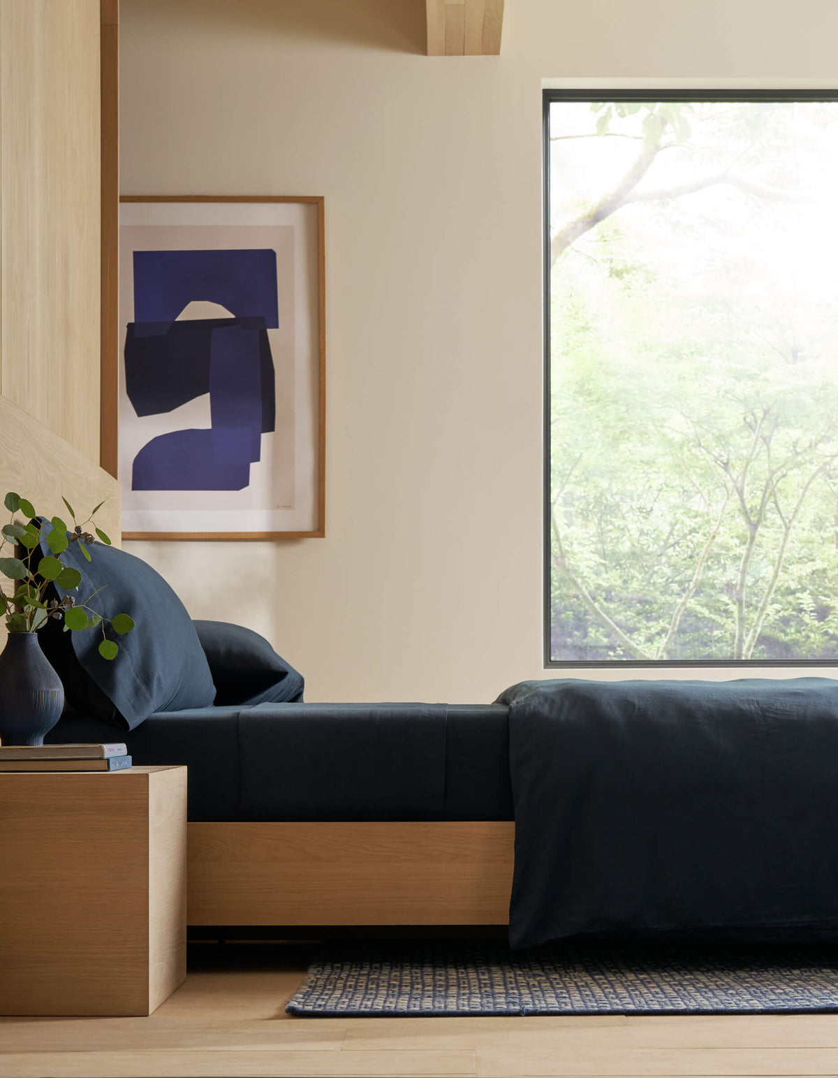 A modern bedroom featuring Cozy Earth's Soft-Wash Cotton Fitted Sheet in dark blue, paired with light wood furniture, a small bedside table adorned with a vase of greenery, abstract blue wall art, and a large window overlooking nature. 