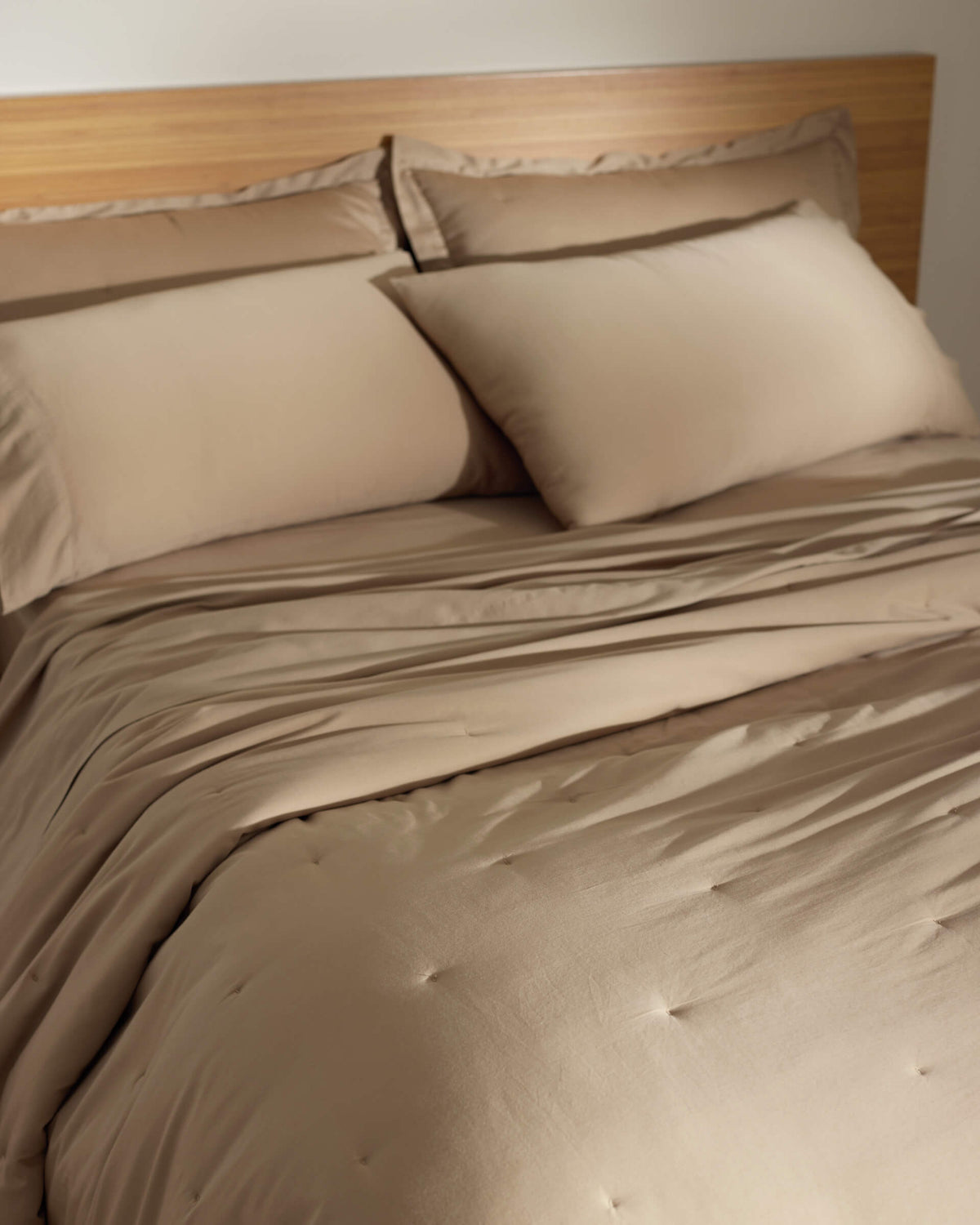 A neatly made bed with the Cozy Earth Soft-Wash Cotton Quilt, featuring beige sheets and pillowcases, is set against a wooden headboard in a softly lit room.