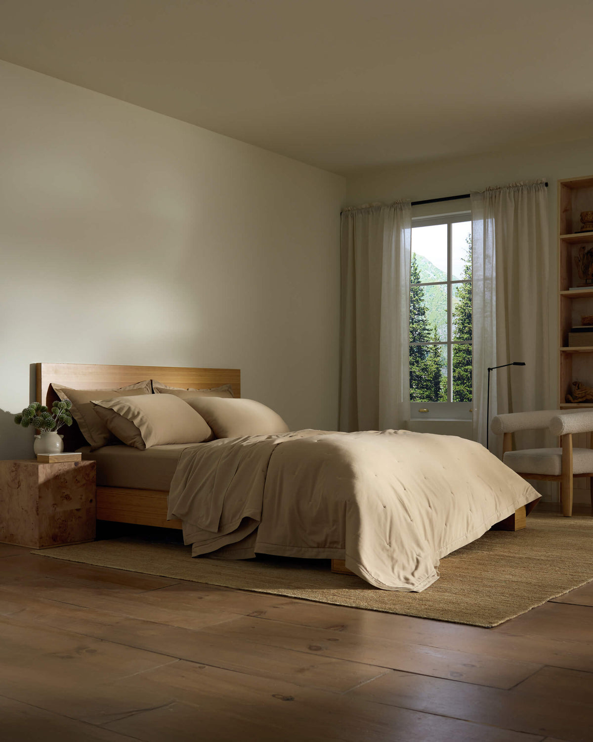 A cozy bedroom features the Cozy Earth Soft-Wash Cotton Quilt on a wooden bed, beige bedding, a bedside table with a plant, sheer-curtained window, cream armchair, wooden shelves, and a woven rug on a wooden floor.