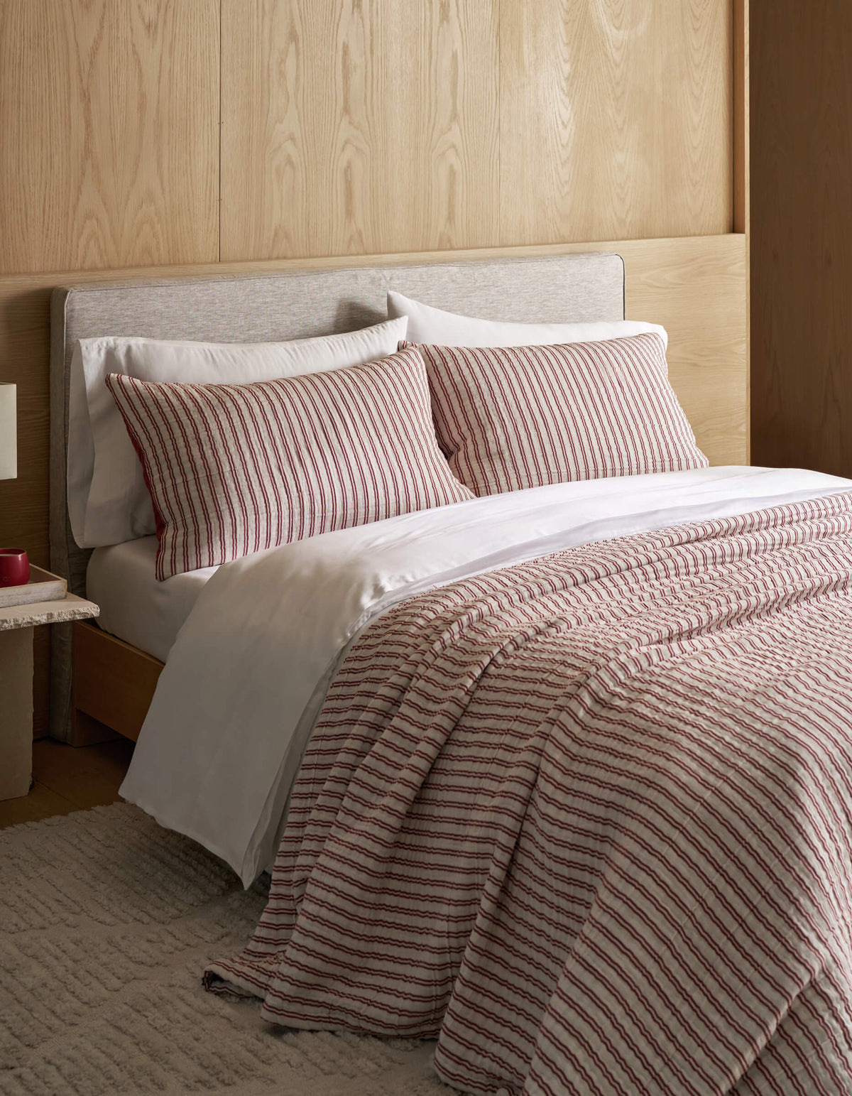 A neatly made bed features the Cozy Earth Waterfront Coverlet Bundle, white sheets, and a striped comforter with matching pillows, set against a light wood headboard and wall, complemented by a beige rug and a small bedside table.