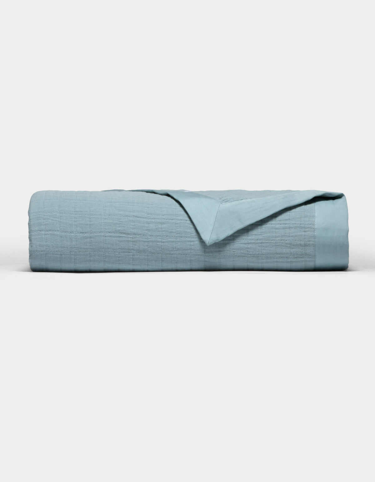 The Cozy Earth Waterfront Grid Matelasse Blanket in light blue is neatly folded against a plain white background, with the top layer turned back to show the inner fabric.