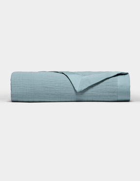 The Cozy Earth Waterfront Grid Matelasse Blanket in light blue is neatly folded against a plain white background, with the top layer turned back to show the inner fabric.|Color:River