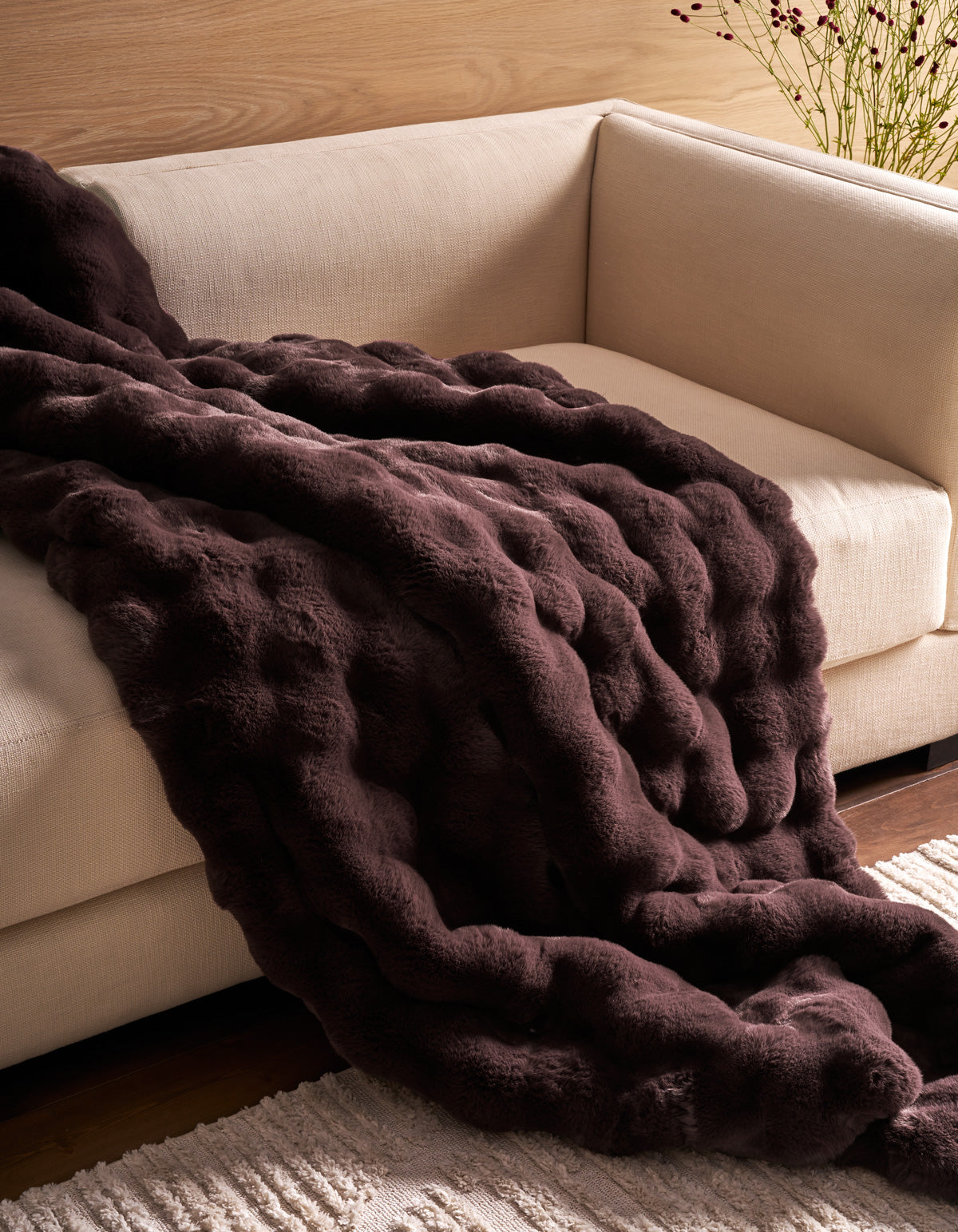 The Cozy Earth Bubble Cuddle Blanket rests atop a light beige sofa, complemented by a cream-colored rug and a wooden wall in the background.|Color:Raisin