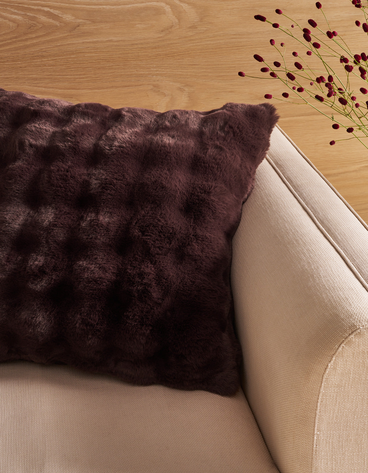 A dark purple textured faux fur throw pillow resting on a beige fabric sofa, with a light wood floor and decorative dried stems in the background. |Color:Raisin |Size:20" x 20"