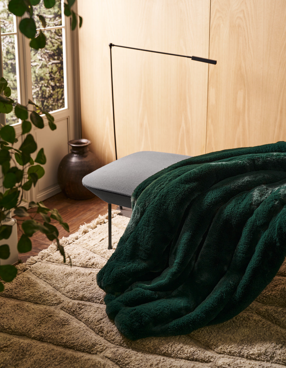The Channel Cuddle Blanket by Cozy Earth, in a rich dark green faux fur, is draped over a gray bench in a cozy room featuring light wood walls, a beige rug, a potted plant, and natural light streaming through the window.|Color:Emerald
