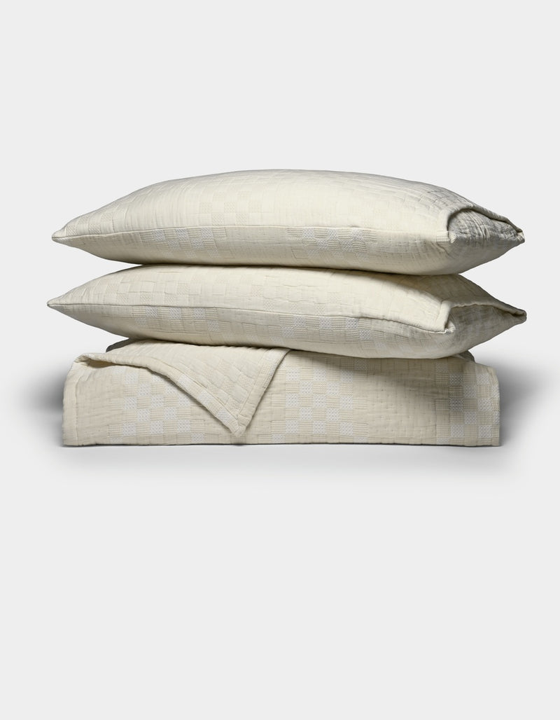 Two cream-colored pillows from the Cozy Earth Sonoma Stitch Quilt Bundle are stacked atop a matching folded blanket, all featuring a subtle checkered pattern, set against a plain white background.