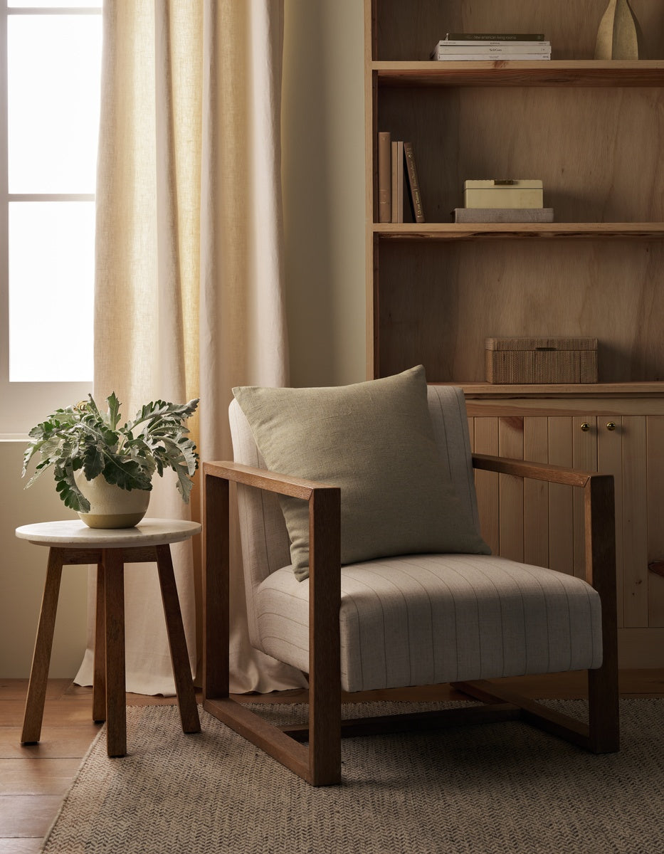 A cozy reading nook with a striped armchair, a neutral Sonoma Throw Pillow by Cozy Earth, a wooden side table holding a potted plant, built-in wooden shelves, a closed cabinet, and sunlight streaming through a window with light curtains.