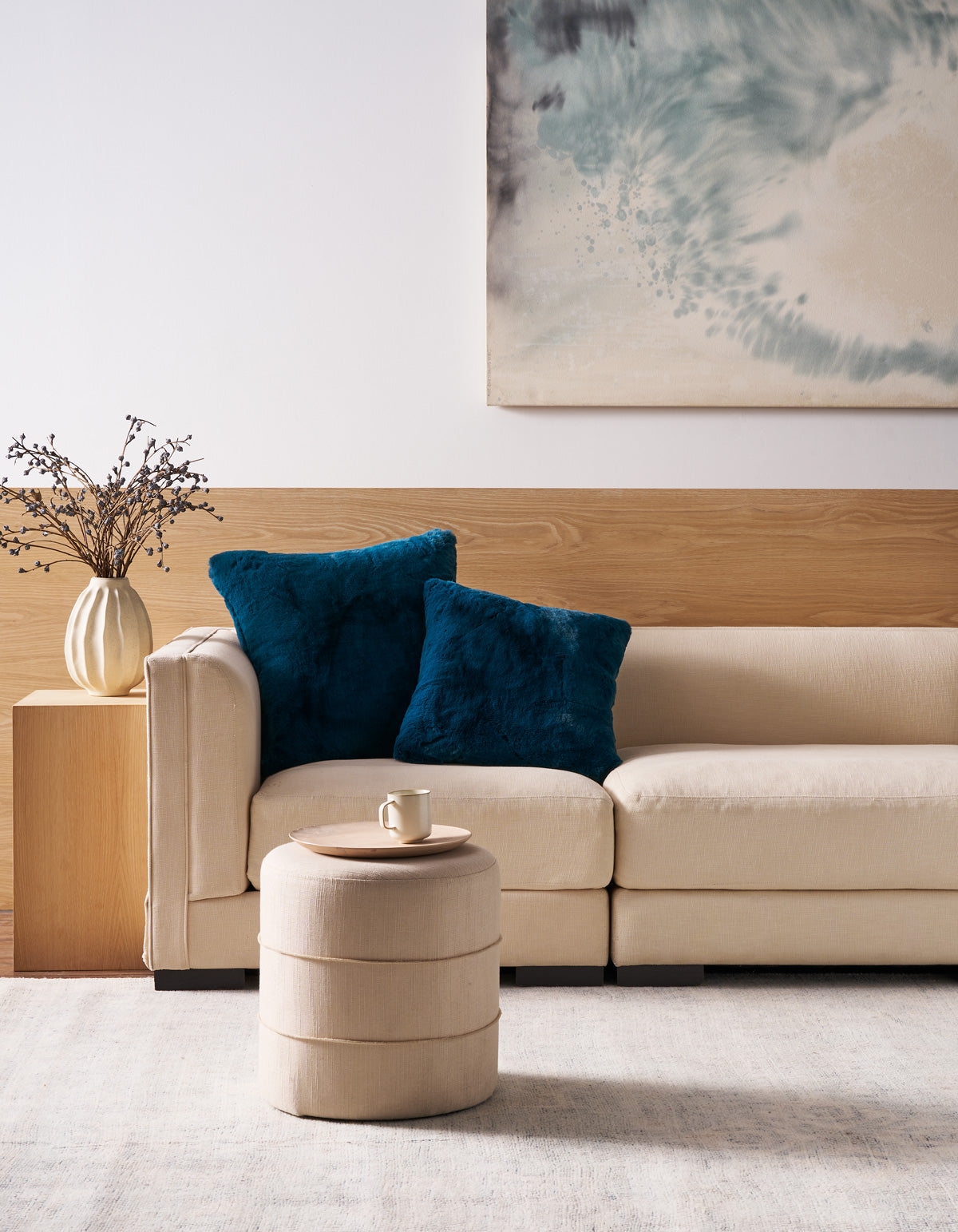 A beige sofa accented with two Cozy Earth Superplush Cuddle Pillows in blue sits against a wooden accent wall. In front, a round beige ottoman holds a cup, while the background is decorated with a vase of branches and a blue abstract painting.|Color:Deep Water |Size:26" x 26"