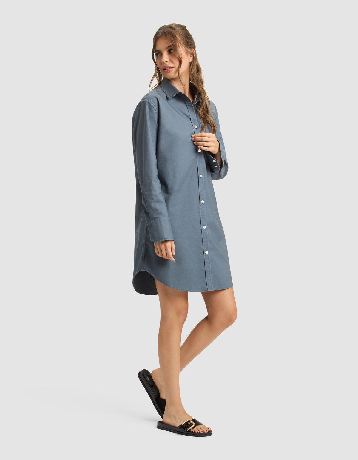 A woman poses in a relaxed way, wearing Cozy Earth's Women's Camden Dress—a loose-fitting, long-sleeve blue shirt dress—paired with black sandals featuring gold accents against a plain light background. 
