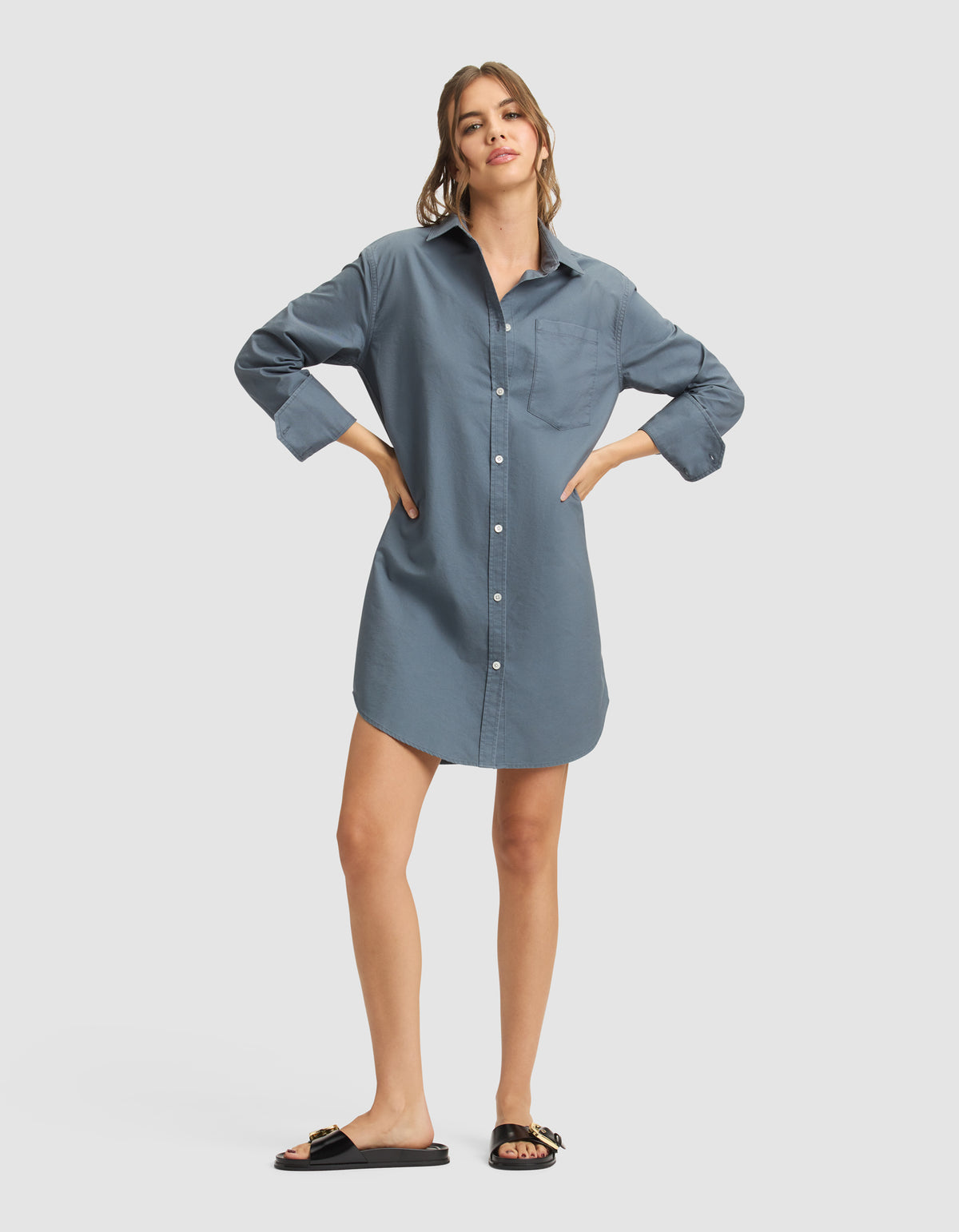 A woman wears the Cozy Earth Women's Camden Dress, a loose-fitting blue button-down shirt-dress, paired with black slide sandals with gold accents. Her long hair is pulled back and she stands against a plain light background. 