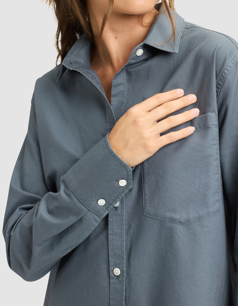A woman wearing the Cozy Earth Women's Camden Dress touches the chest pocket with her right hand. Her face is partially out of frame, and the background is plain light gray.