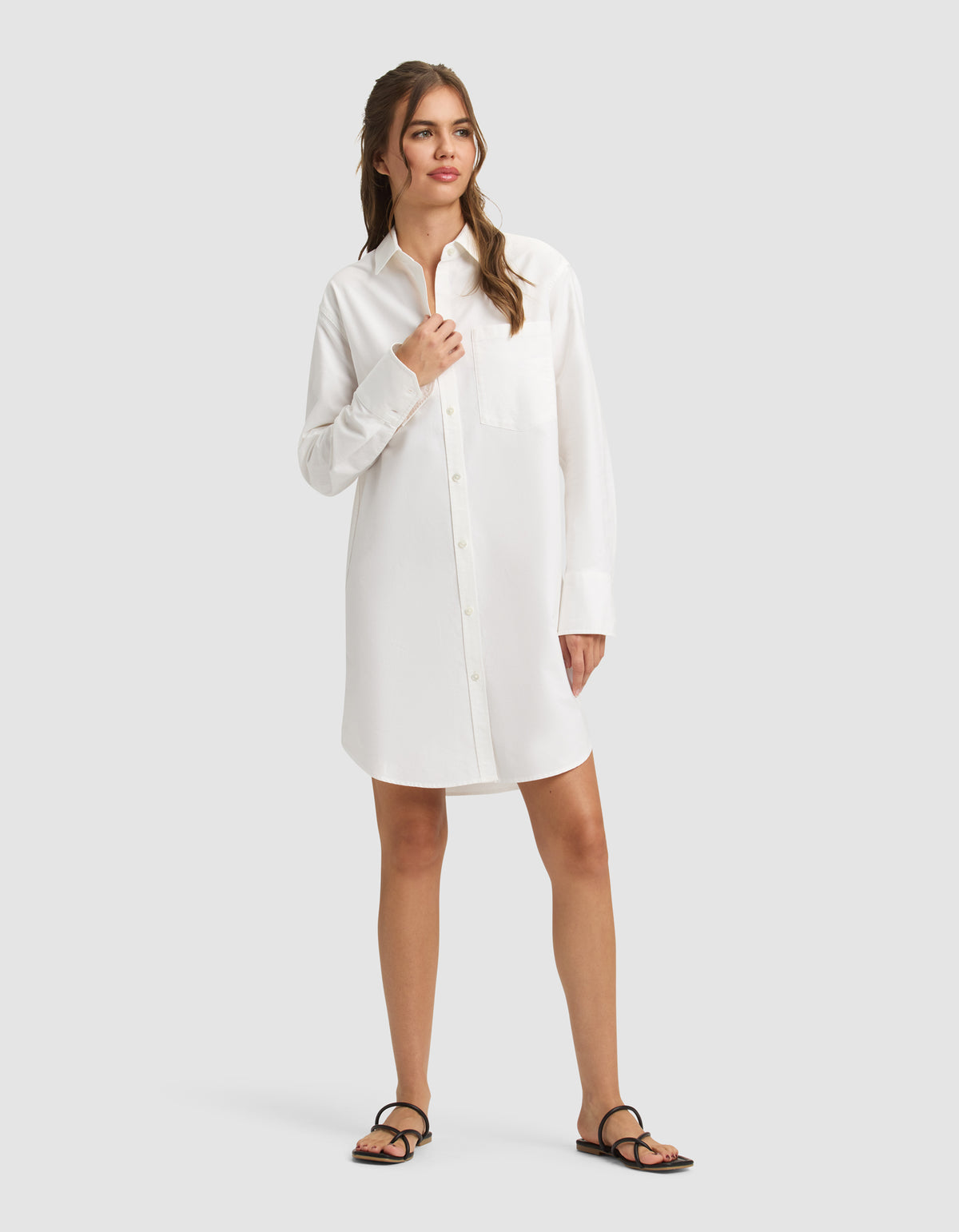 A woman with light brown hair stands against a plain background wearing the Cozy Earth Women's Camden Dress, a long-sleeved white button-up shirt dress with a front pocket, paired with black sandals. She gazes to the side with a relaxed expression. |Color:White