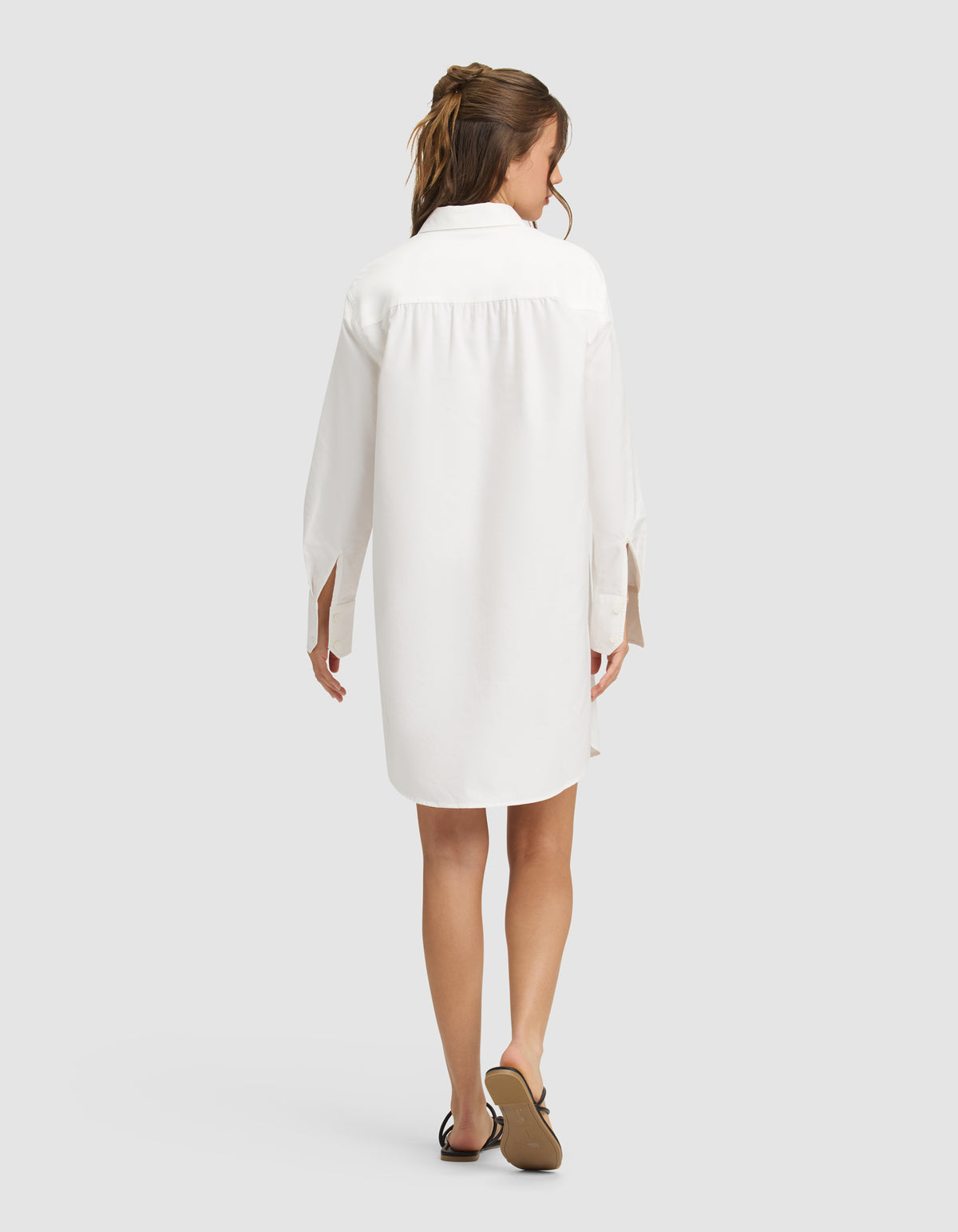 A woman with long brown hair in a loose ponytail is shown from behind, wearing the Cozy Earth Women's Camden Dress—a white, long-sleeve, knee-length shirt dress—with black sandals, standing against a plain light gray background. 