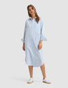 A woman wears the Cozy Earth Women's Camden Midi Dress, a loose light blue, long-sleeve, button-down shirt dress with subtle stripes. She pairs it with white sneakers and smiles slightly, posing with one hand by her side. |Color:Blue Oxford Stripe