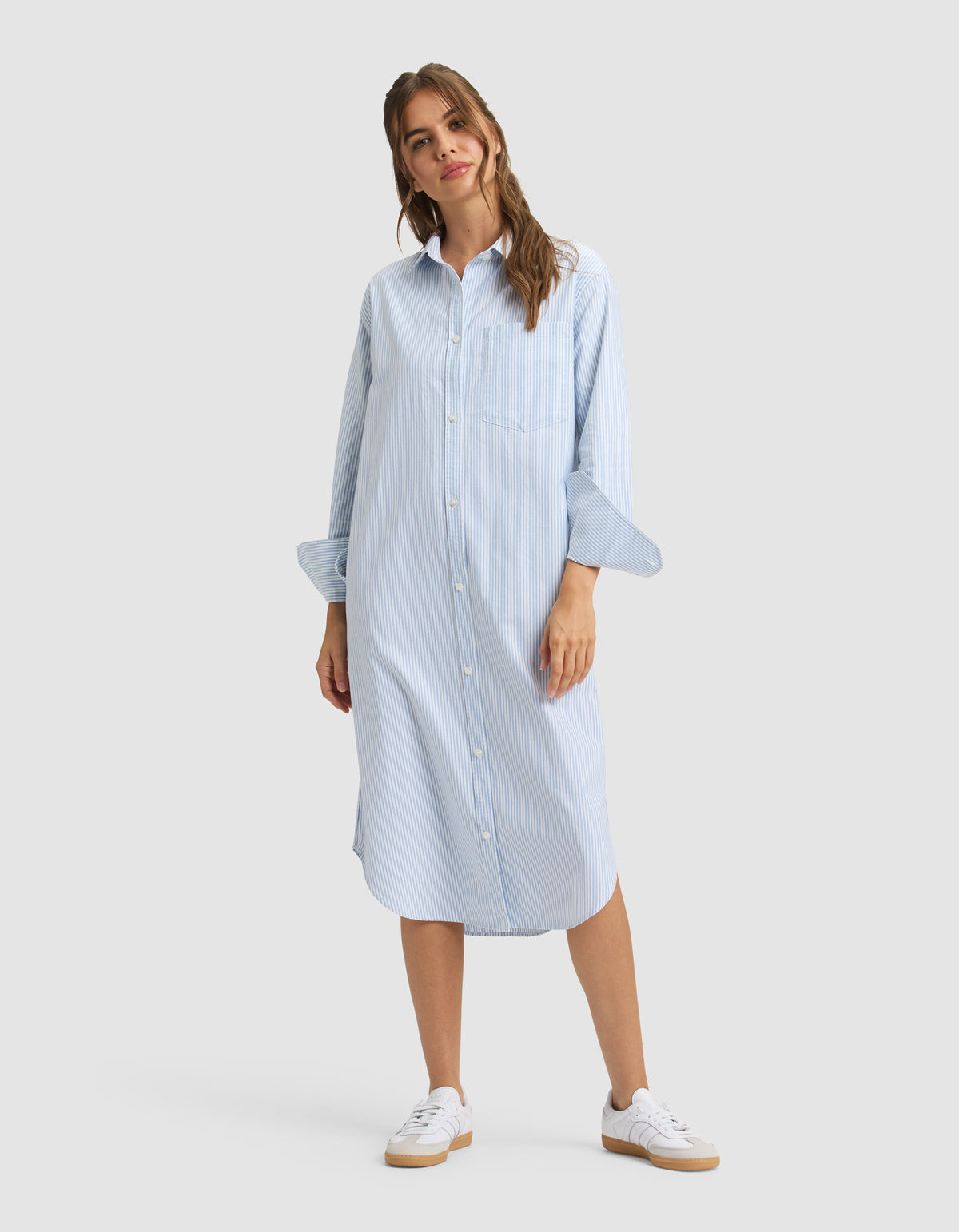 A woman wears the Cozy Earth Women's Camden Midi Dress, a loose light blue, long-sleeve, button-down shirt dress with subtle stripes. She pairs it with white sneakers and smiles slightly, posing with one hand by her side. 