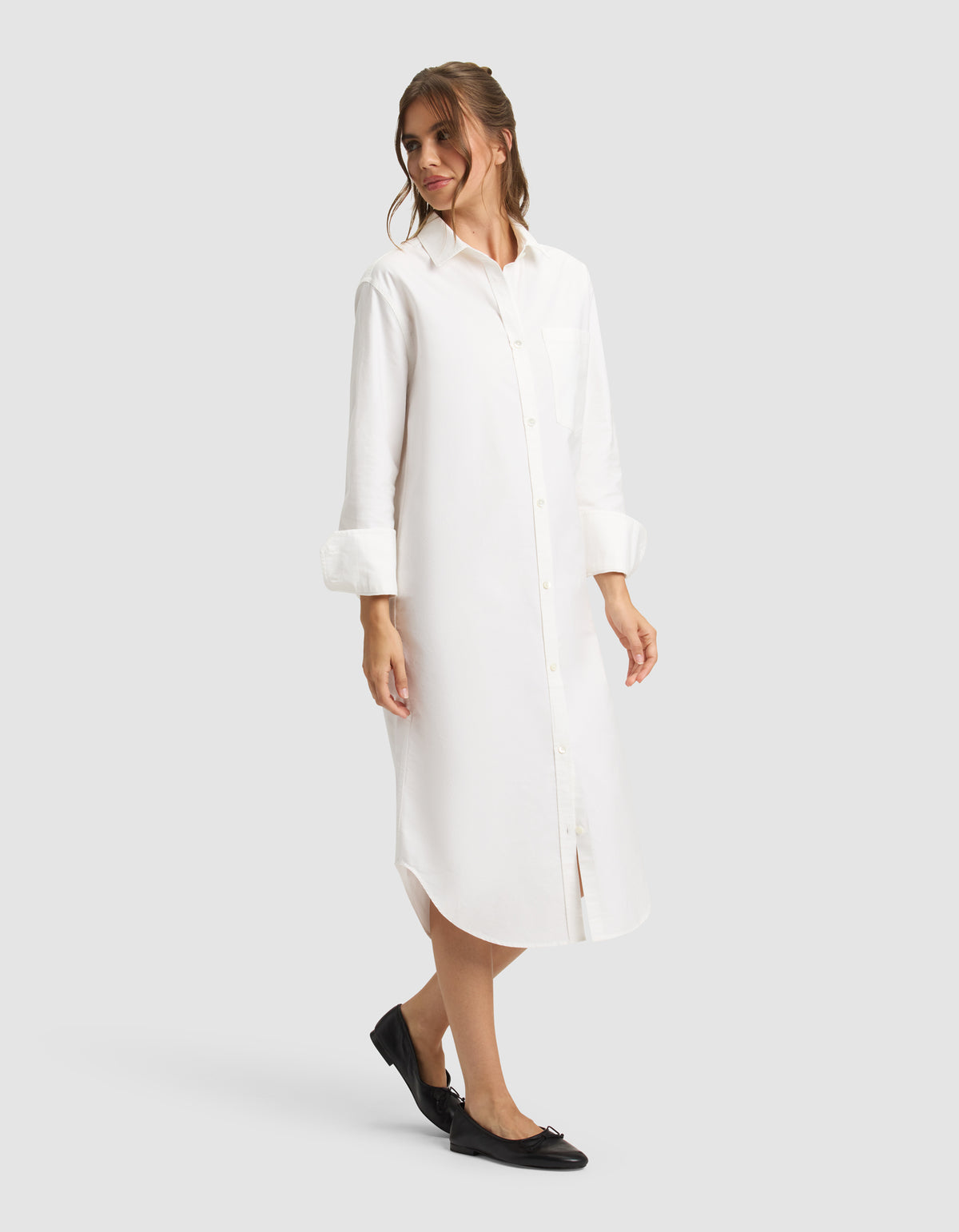 A woman models the Cozy Earth Women's Camden Midi Dress—a long white button-down shirt dress with rolled-up sleeves—paired with black flats, standing against a plain light gray backdrop. 