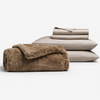 The Cozy Night In Bundle by Cozy Earth features a folded brown plush blanket, two stacked beige pillows, and a neatly folded beige sheet, all displayed against a white background.