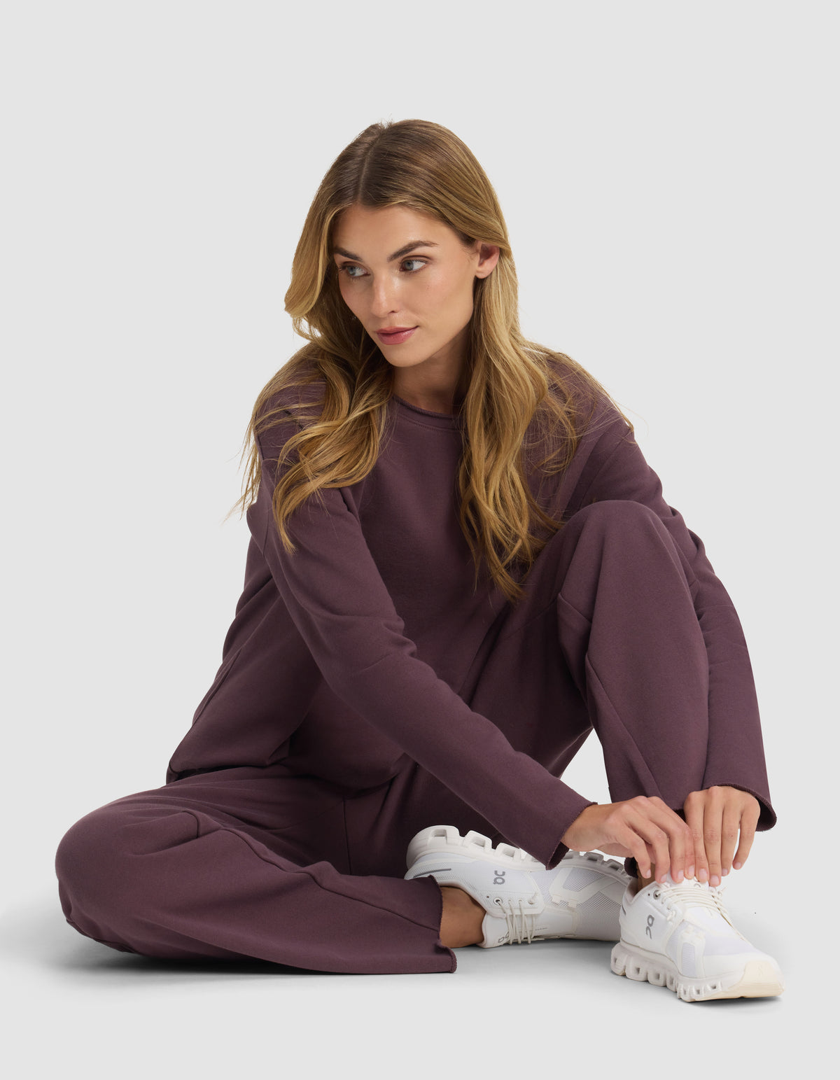 A woman with long blonde hair sits on the floor wearing a loose-fitting mauve Chelsea Pant by Cozy Earth and white sneakers, looking to her right while adjusting her shoe against a plain light background.