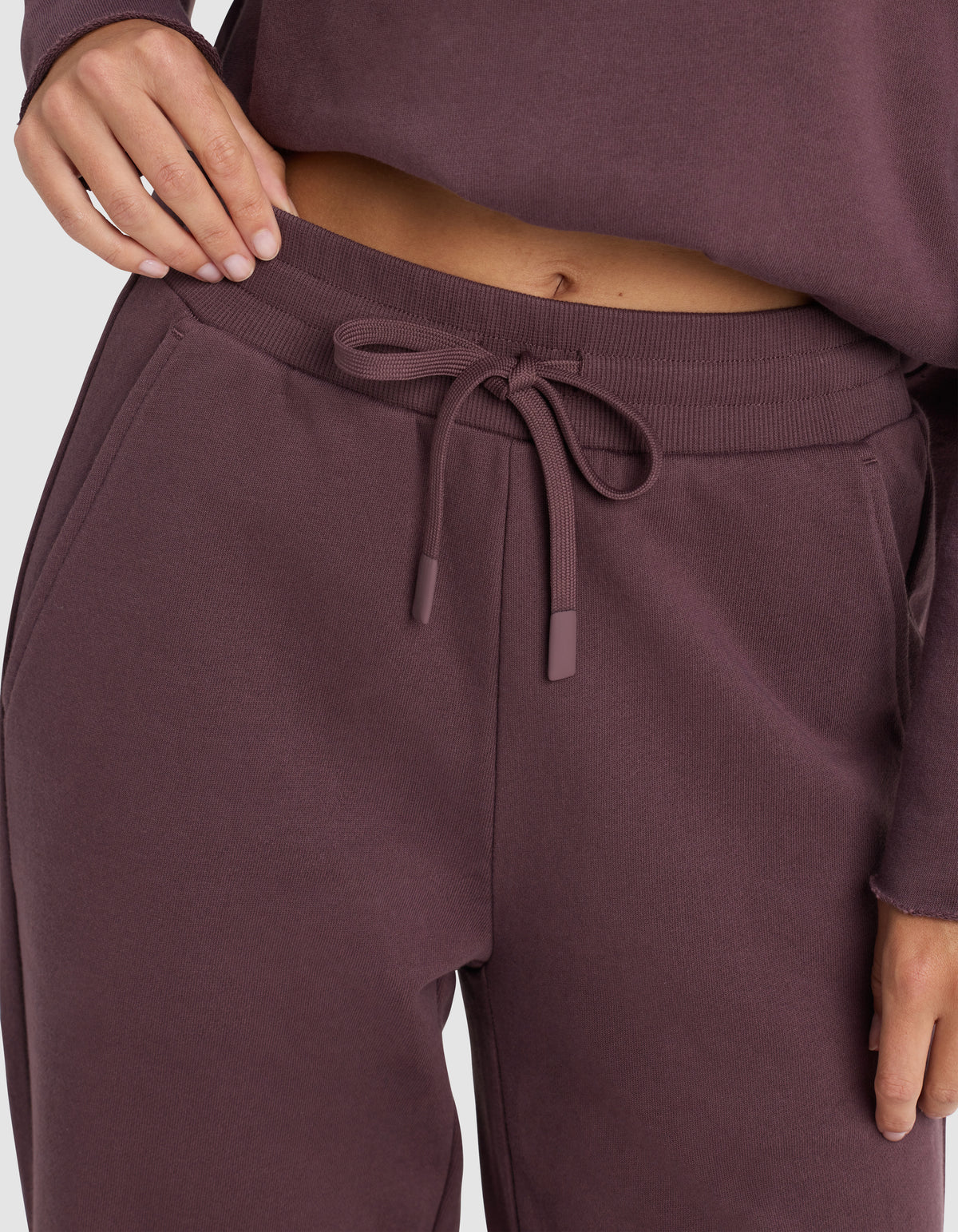 A person wears Cozy Earth's Chelsea Pant, a soft and comfortable drawstring sweatpant, in matching mauve or dark purple, with their hand resting on the waistband.