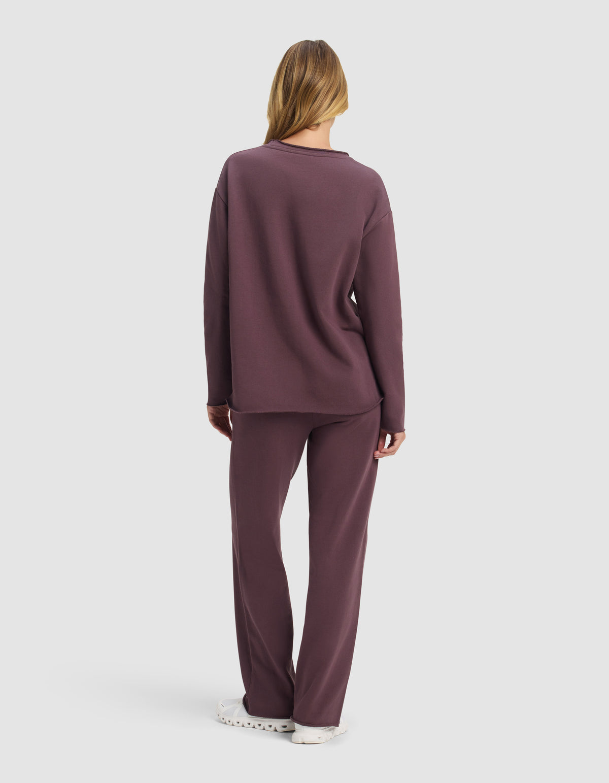 A woman with long hair, seen from behind, wears the Cozy Earth Women's Chelsea Set—a loose-fitting, long-sleeve top and matching pants in muted purple—paired with white shoes against a plain white background.