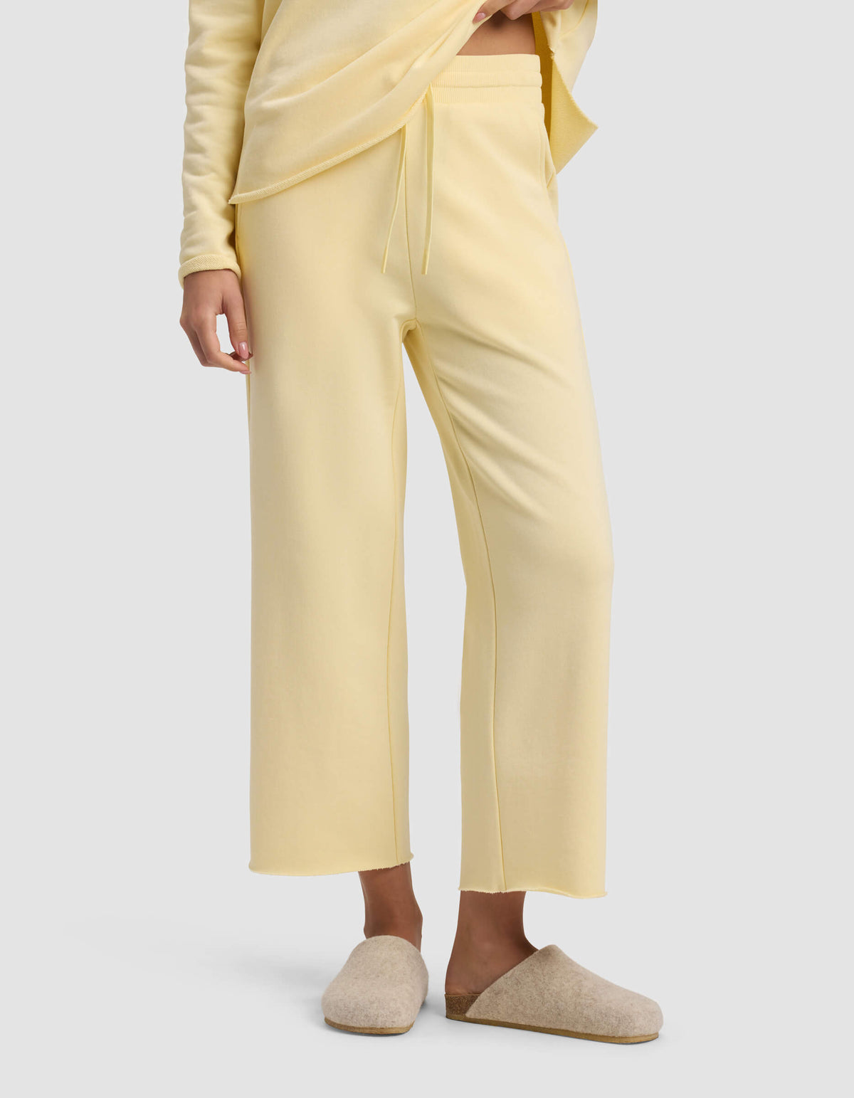A person is wearing Cozy Earth's Women's Chelsea Cropped Pant in light yellow with an elastic waistband and drawstring, paired with a matching top and beige slippers, standing against a plain background.