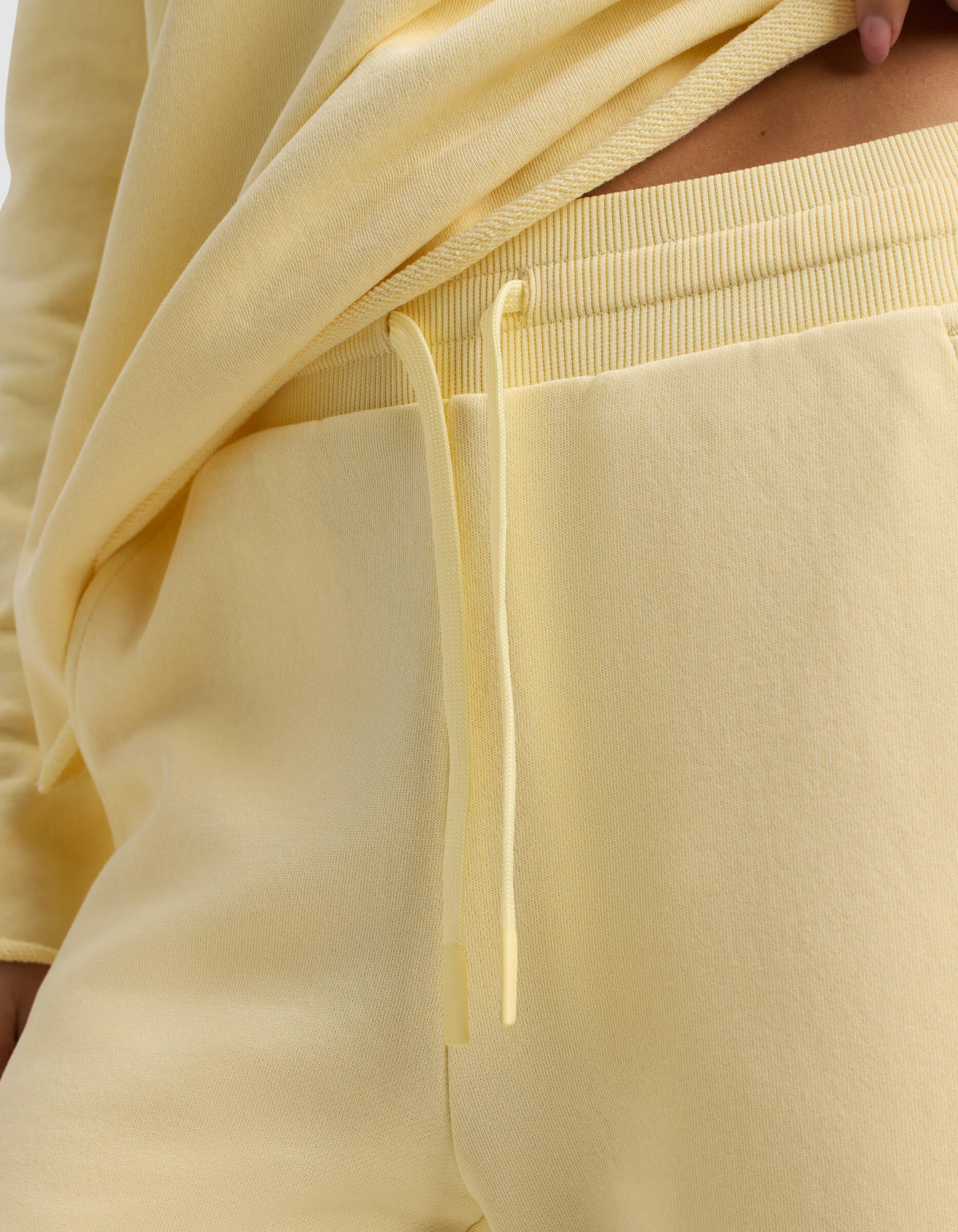 Close-up of a person wearing the Women's Chelsea Cropped Pant by Cozy Earth in light yellow, featuring a matching drawstring and paired with a yellow sweatshirt slightly lifted to reveal the waistband.
