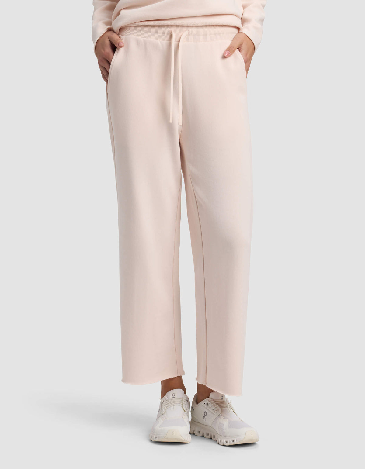 A person wears Cozy Earth Women's Chelsea Cropped Pant in light pink, styled with white sneakers and hands in pockets, against a plain light background.|Color:Petal