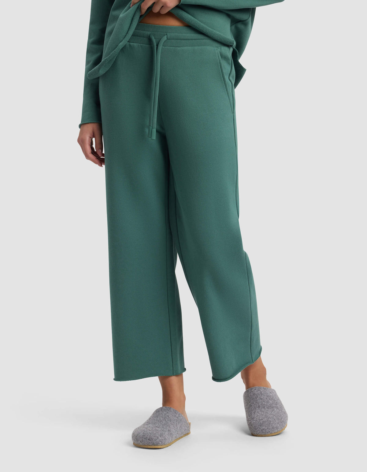 A person wears the Cozy Earth Women's Chelsea Cropped Pant in green with a drawstring, a matching top partly visible, and gray slippers, standing against a plain light gray background.|Color:Sea Green
