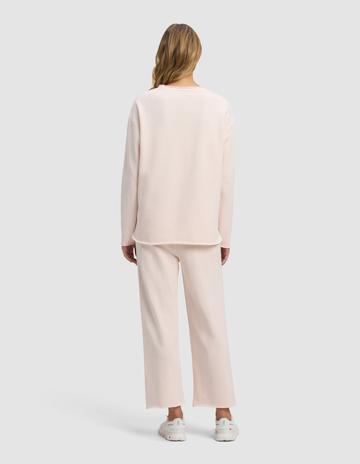 A woman with long, wavy blonde hair stands facing away, wearing the Cozy Earth Women's Chelsea Cropped Set in light pink with white sneakers, set against a plain gray background.