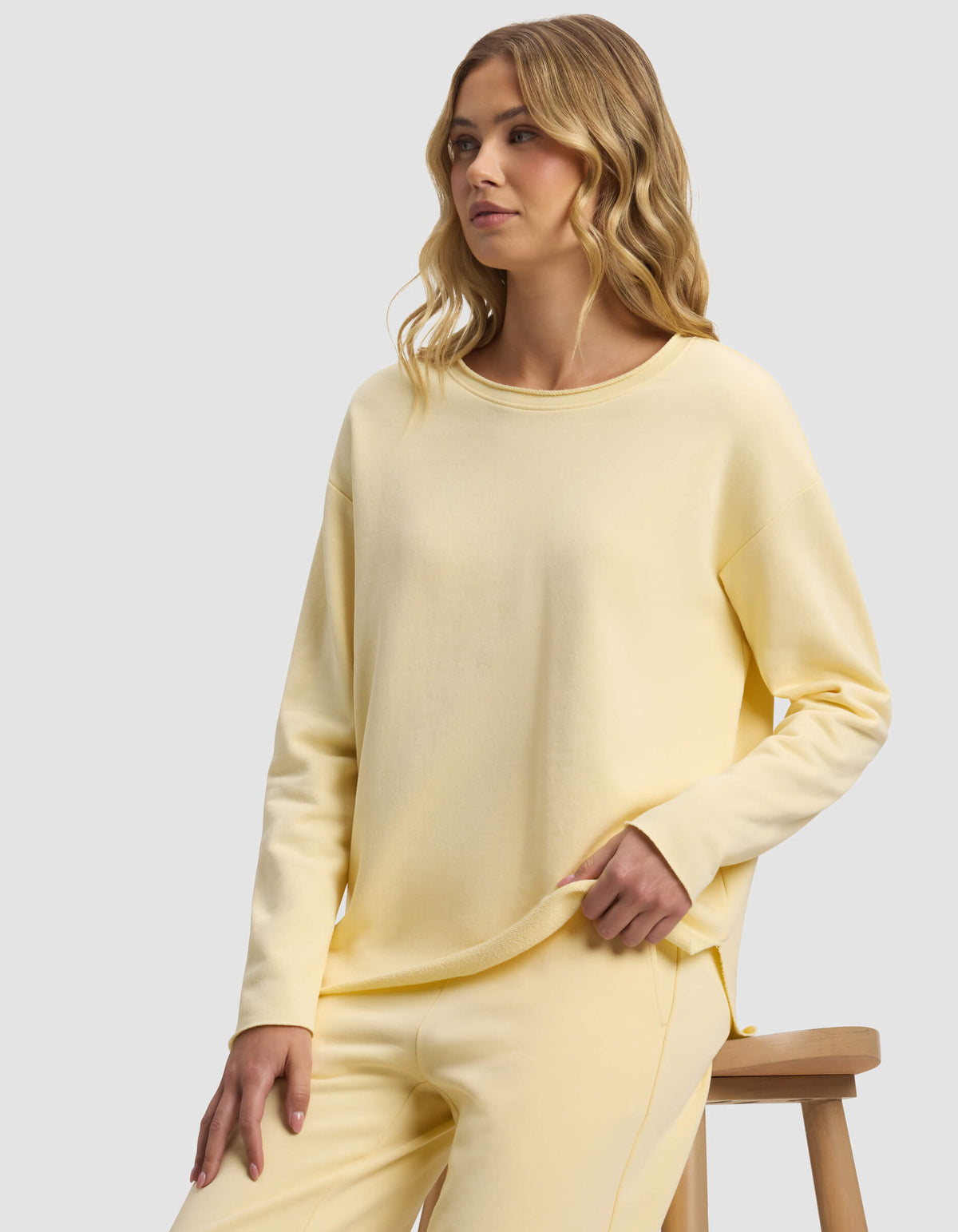 A woman with wavy blonde hair models the Cozy Earth Women's Chelsea Pullover in pastel yellow, paired with matching pants, as she sits on a wooden stool against a plain light gray background.