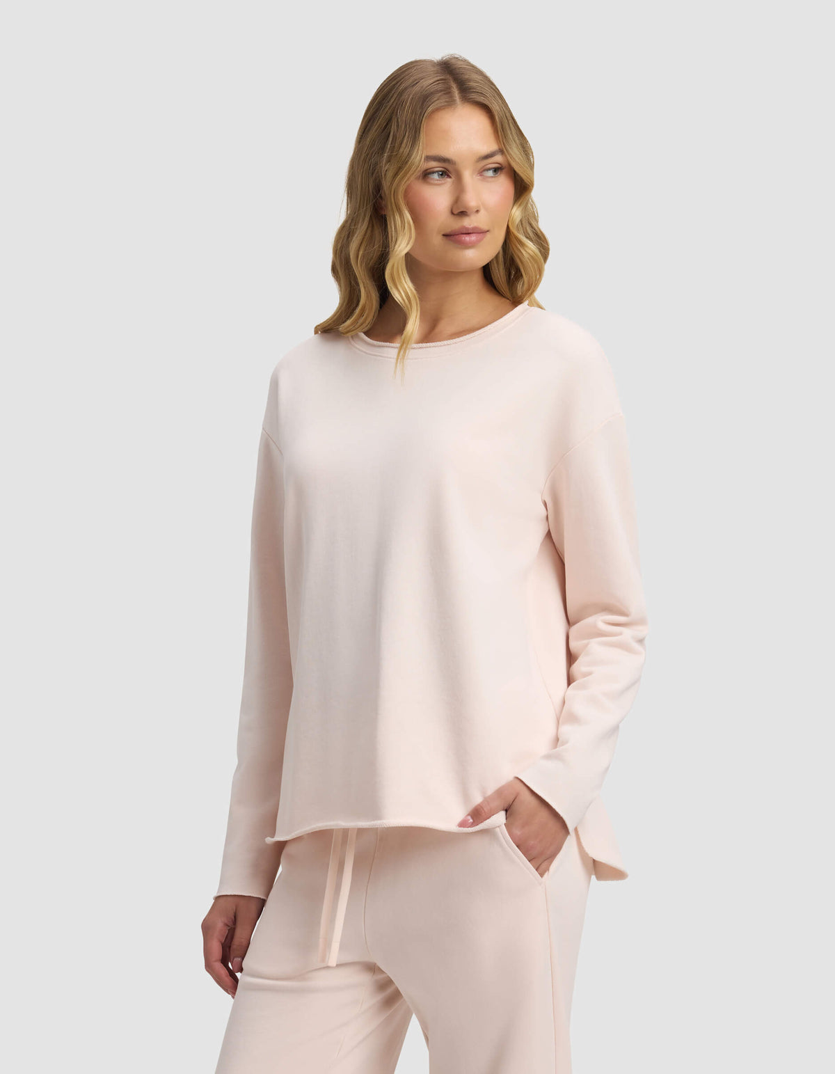 A woman with wavy blonde hair models the Cozy Earth Women's Chelsea Pullover in pale pink, paired with matching lounge pants, standing with one hand in her pocket against a plain light gray background.