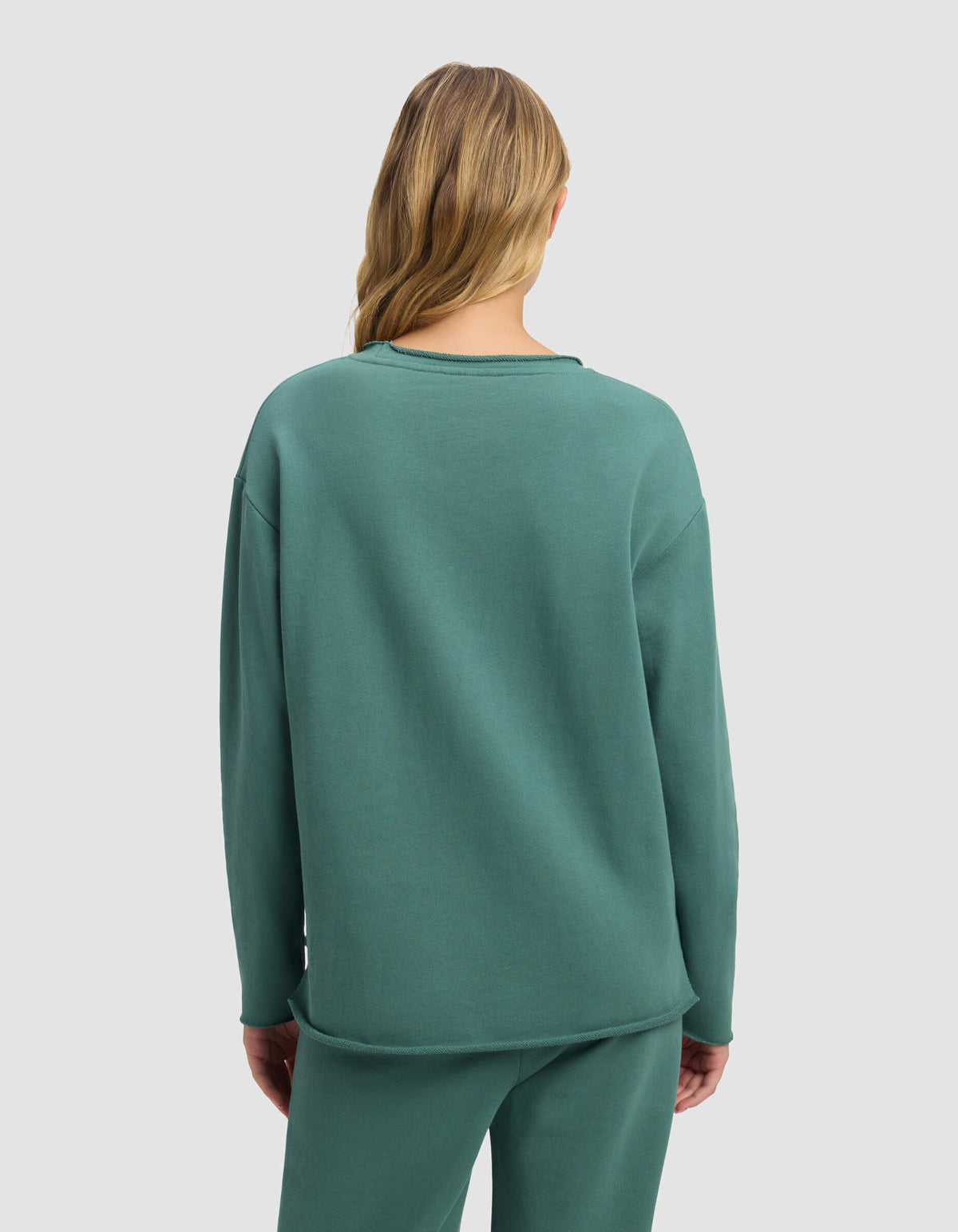 A woman with long, wavy blonde hair is seen from behind wearing the Cozy Earth Women's Chelsea Pullover in teal, paired with matching pants, set against a plain light gray background.