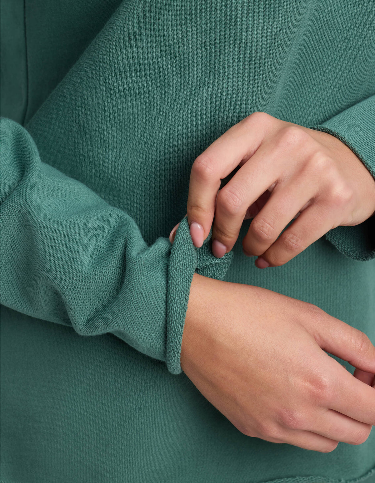 Close-up of a woman wearing the Cozy Earth Women's Chelsea Pullover in green, adjusting the cuff on her left sleeve with her right hand. The out-of-focus background highlights the hands and textured fabric.