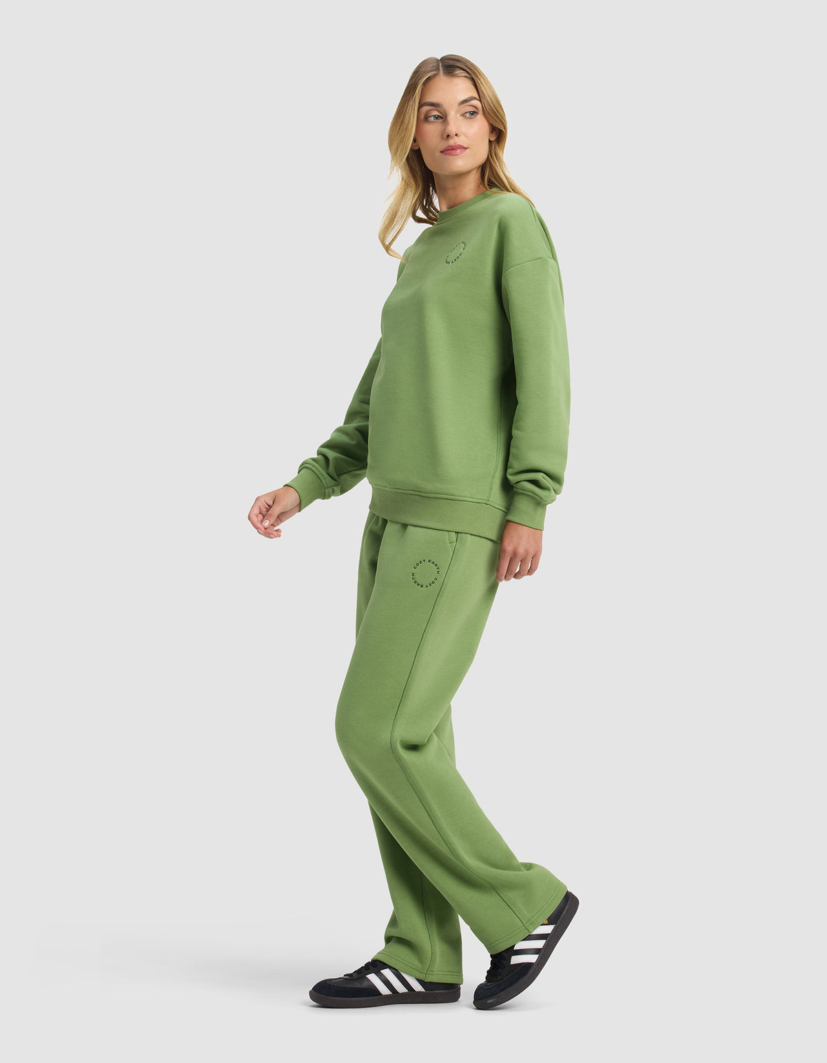 A woman stands sideways wearing the Cozy Earth Women's Cityscape Crewneck & Wide Leg Pant Set in green with black and white sneakers. The loose, casual outfit is shown against a plain white background as she looks over her shoulder.