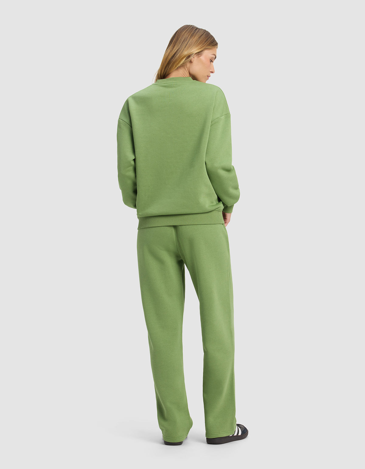A woman faces away, wearing the Cozy Earth Women's Cityscape Crewneck & Wide Leg Pant Set in light green with black and white sneakers, standing against a plain light gray background.