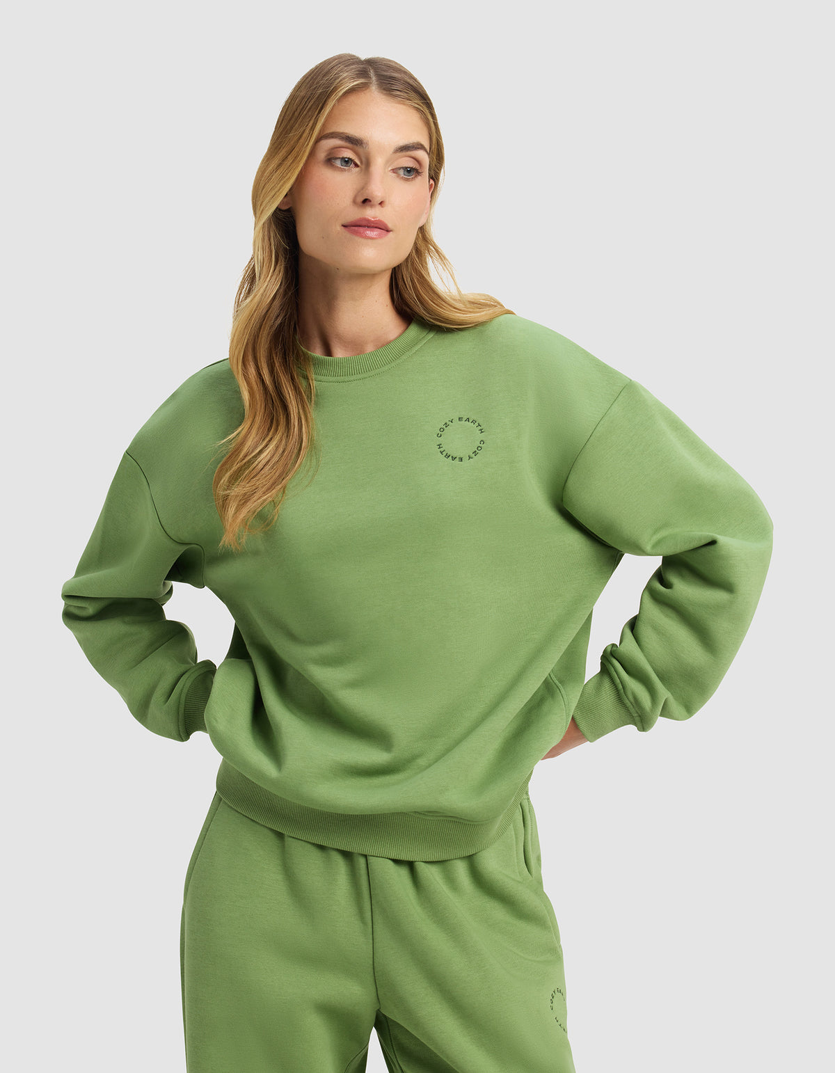 A woman with long blonde hair stands with her hands on her hips against a plain light gray background, wearing the Cozy Earth Women's Cityscape Crewneck in a loose-fitting, light green style with matching sweatpants.