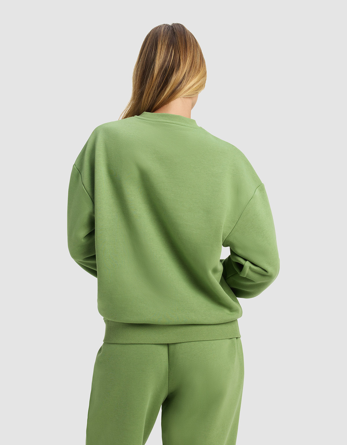 A woman with long, light brown hair is seen from behind wearing the Cozy Earth Women's Cityscape Crewneck in light green, paired with matching sweatpants, standing against a plain light gray background.