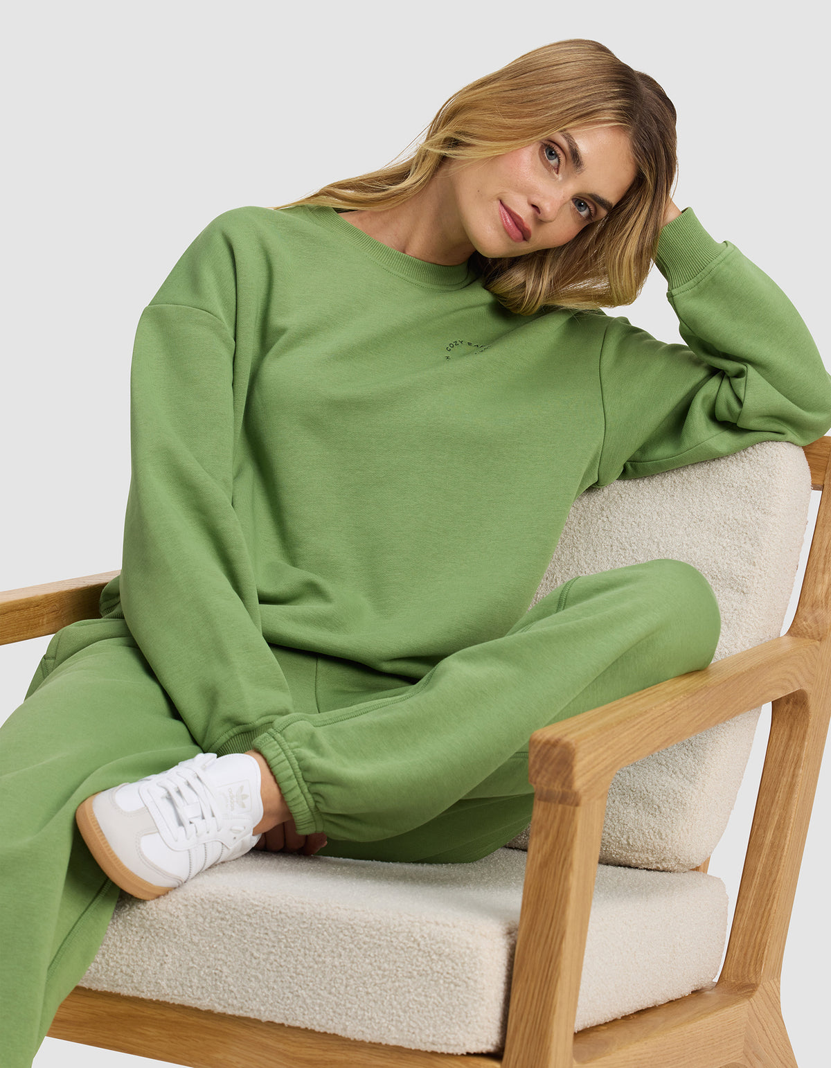 A woman wears the Cozy Earth Women's Cityscape Crewneck, lounging casually on a cushioned wooden chair and looking relaxed in her stylish, comfortable outfit.