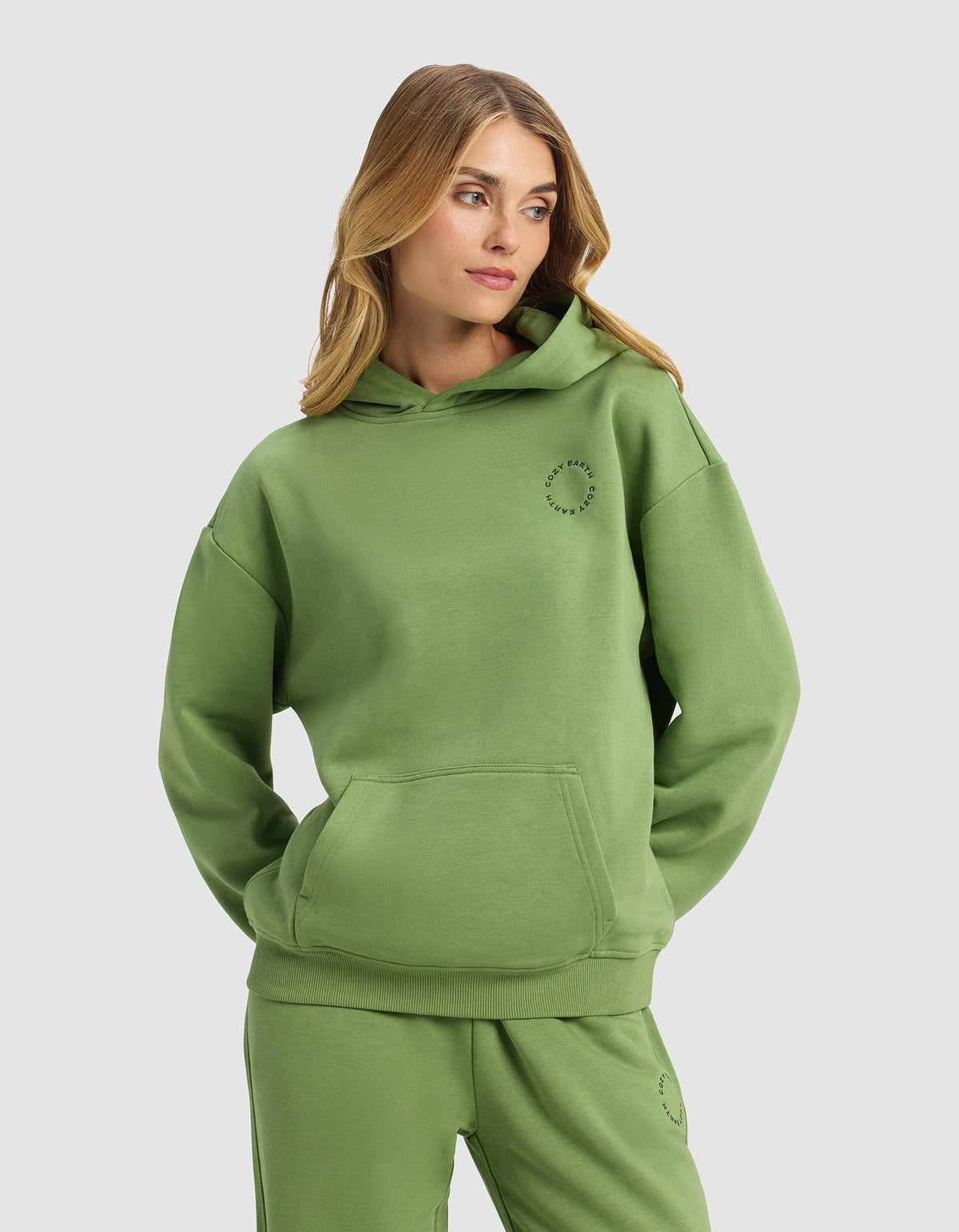A woman with long blonde hair models the Cozy Earth Women's Cityscape Hoodie in green, paired with matching sweatpants, standing with hands in her hoodie pocket against a plain light gray background.